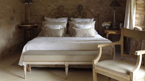 A bedroom with a large bed, two bedside tables, a chair, and a stone wall.