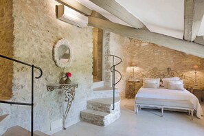 A bedroom with a stone wall, a bed, a nightstand, a mirror, and a staircase.