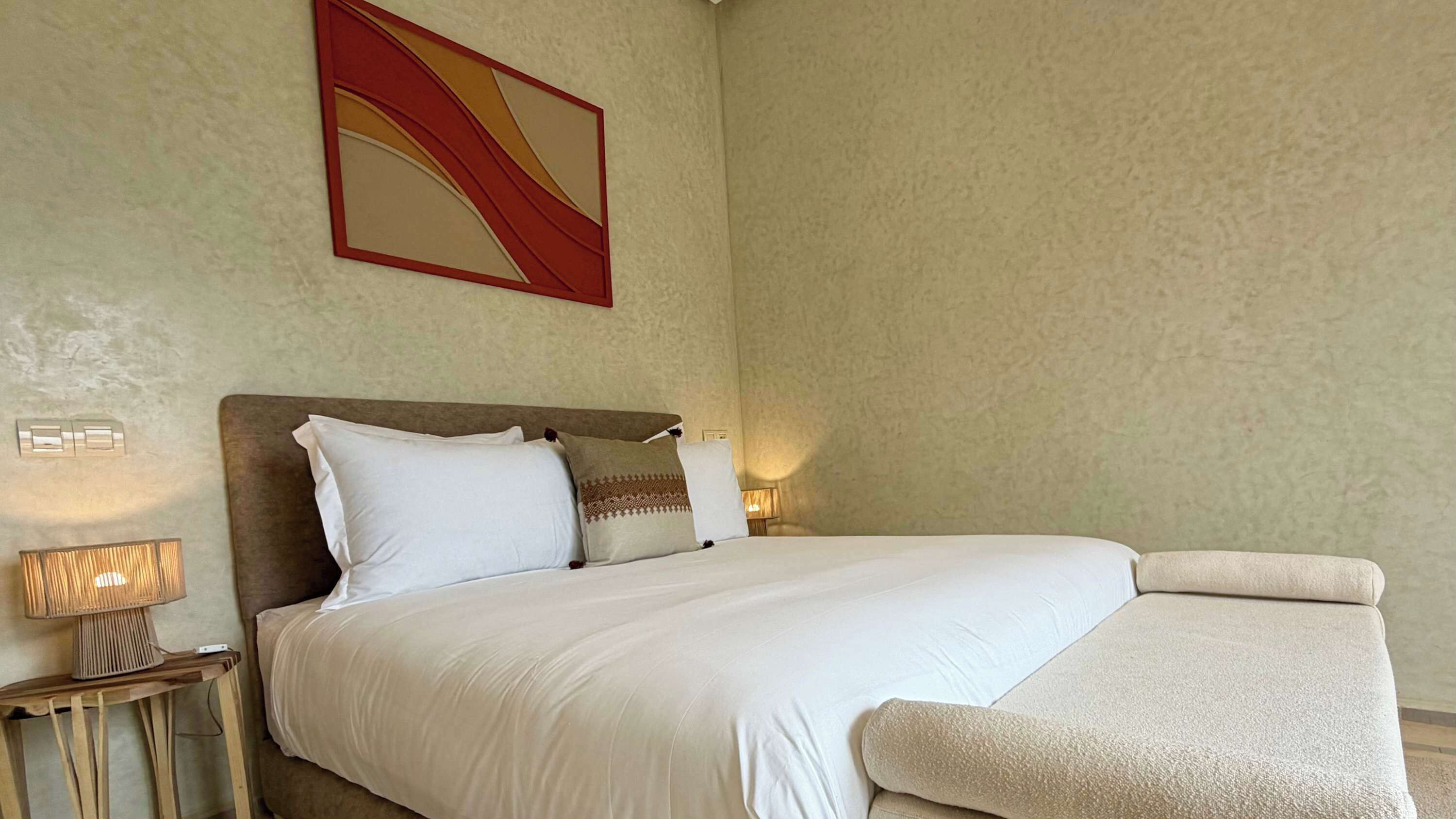 A hotel room with a large bed, a headboard, two bedside lamps, a wall-mounted artwork, and a small table with a lamp.