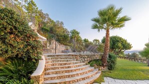 Un escalier en pierre mène à une zone délimitée par une grille, où se trouvent un palmier et une végétation luxuriante.