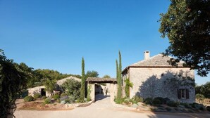Une maison en pierre avec une allée en gravier, entourée d’arbres et de végétation.