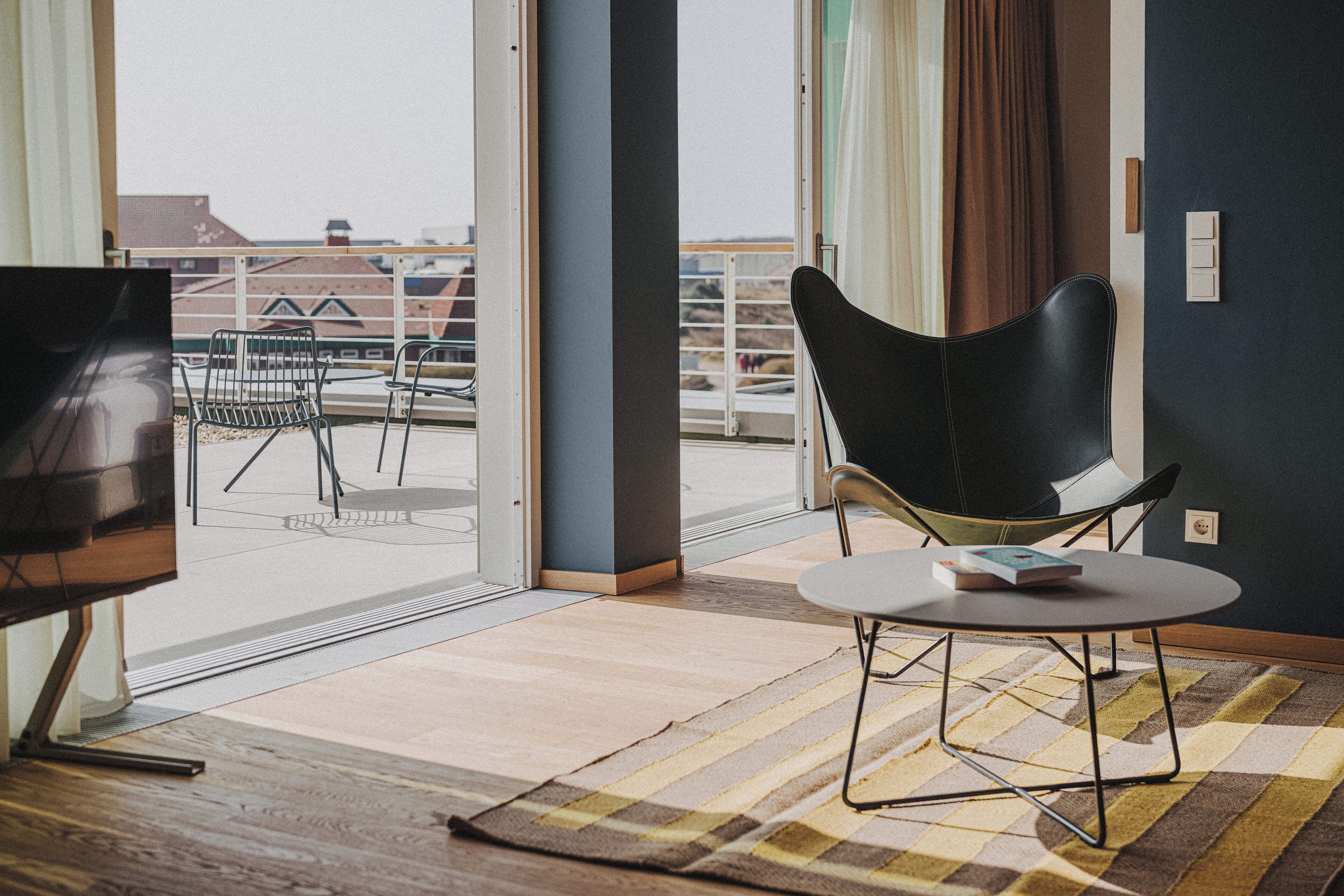 A modern hotel room with a balcony, a flat-screen TV, a dining table, and a black chair.