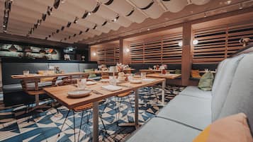 Ein modernes Restaurant mit einem langen Holztisch, Korbstühlen und einem gemusterten Boden.