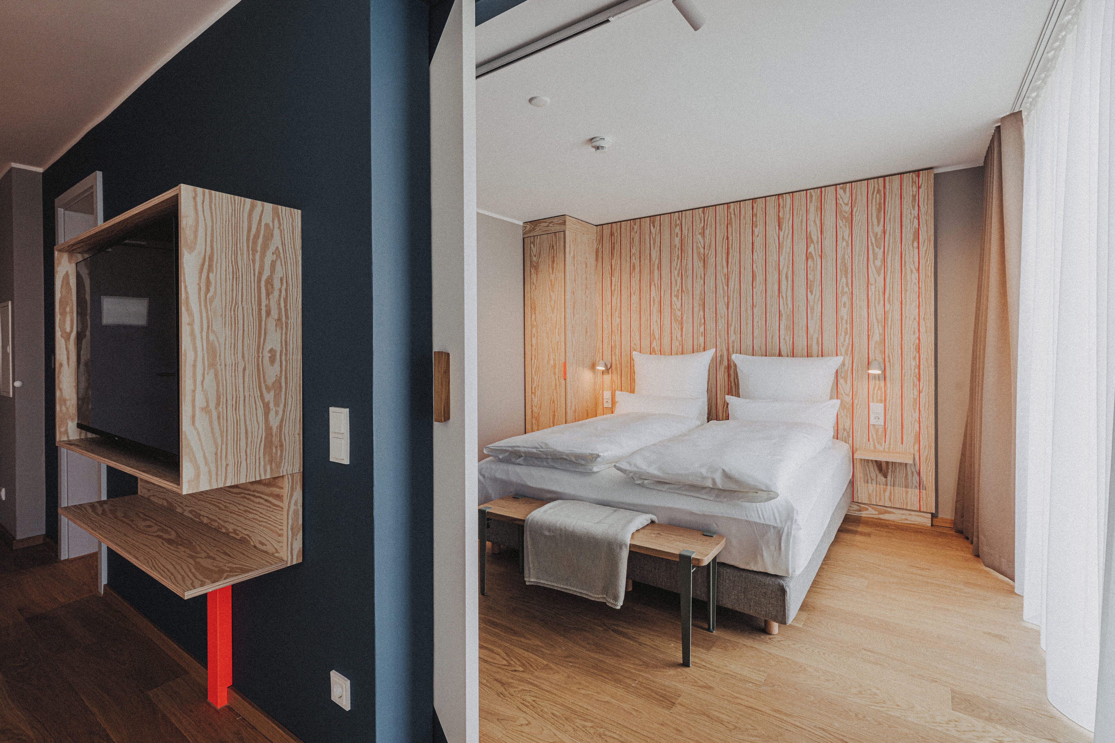 A modern hotel room with a bed, a bench, a TV mounted on a wooden shelf, and a wooden panel wall.