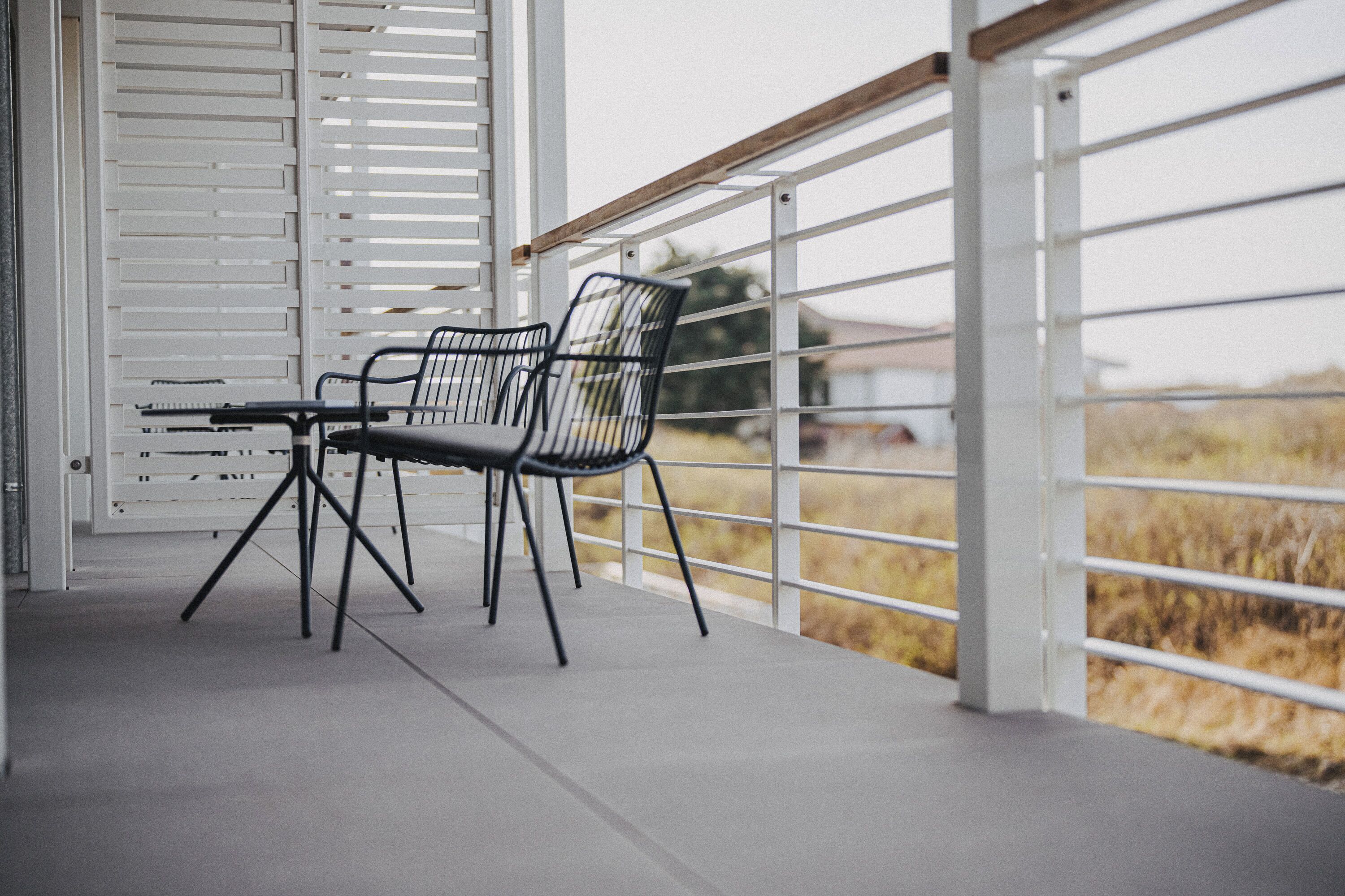 Ein Balkon mit zwei Stühlen, einem Tisch, weißen Geländern und Blick auf ein Feld.