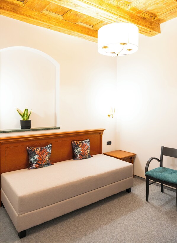 A bedroom with a beige sofa, a wooden headboard, a small bedside table, a green chair, a potted plant, and a wall-mounted light fixture. - Epic Rooms (Brasov)