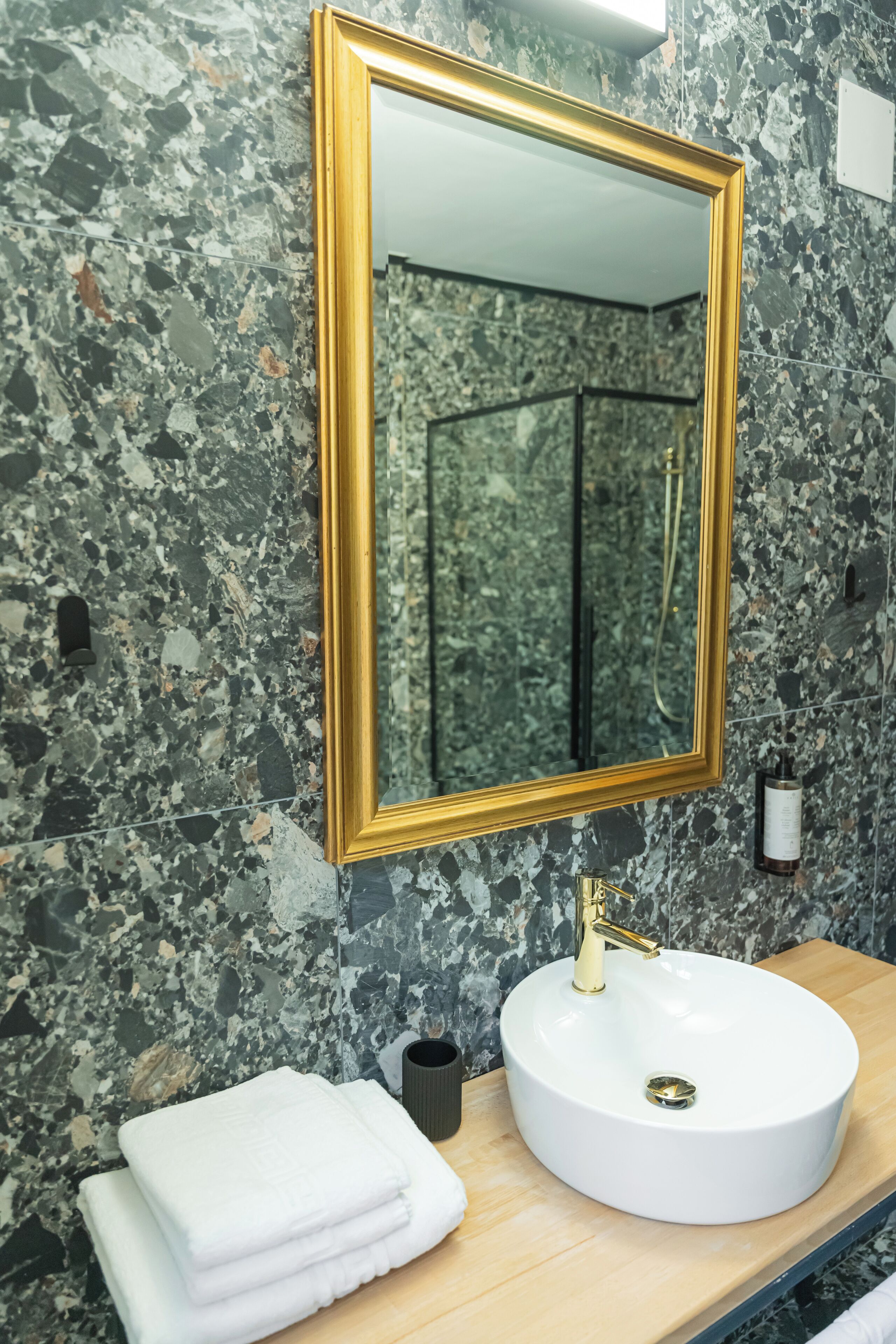 A bathroom with a marble wall, a gold-framed mirror, a white sink, and a towel stack.