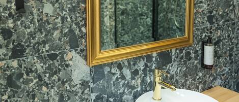 A bathroom with a marble wall, a gold-framed mirror, a white sink, and a towel stack.