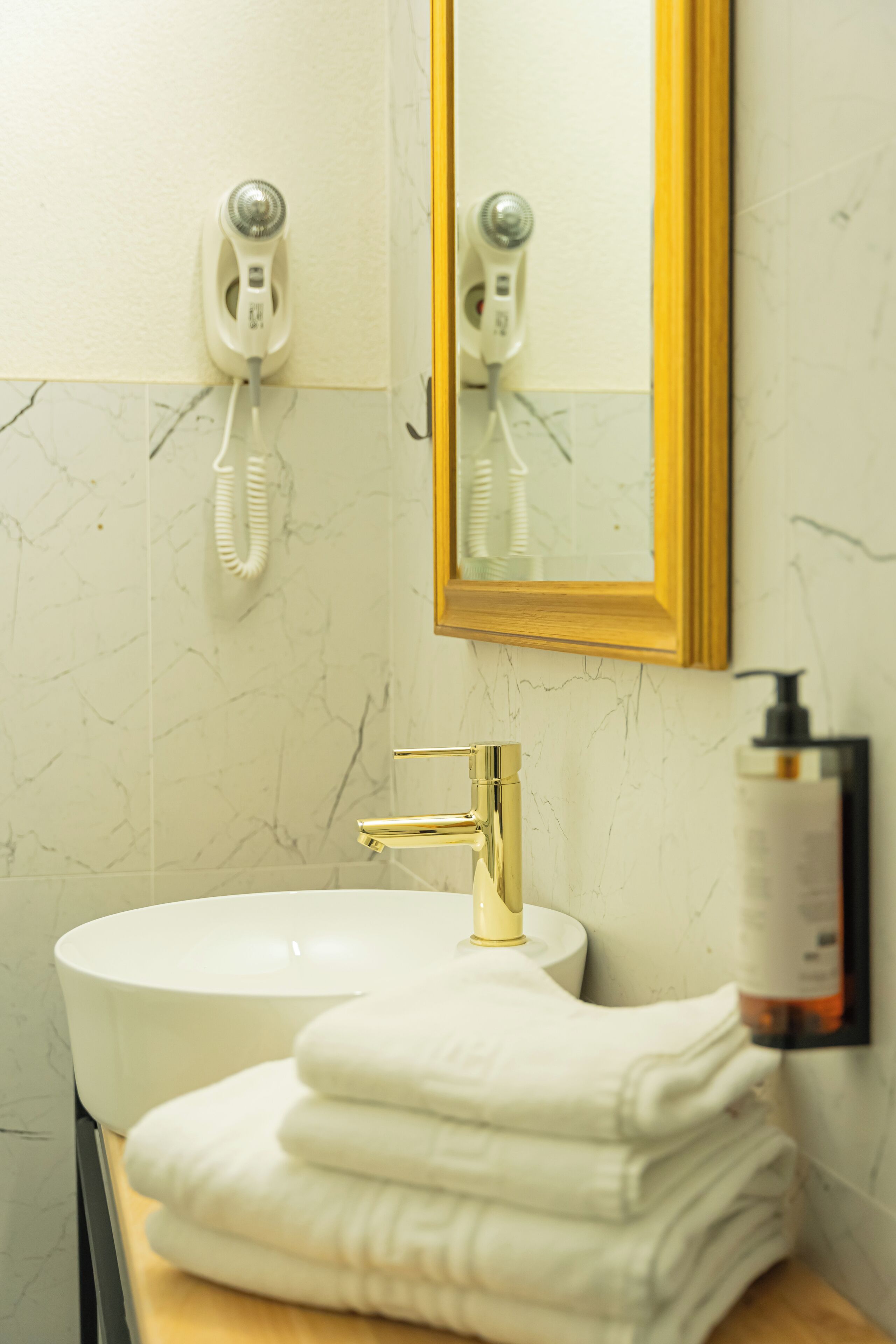 A bathroom with a white sink, gold faucet, and a mirror with a gold frame. There are two toothbrushes mounted on the wall and a bottle of shampoo on the counter.
