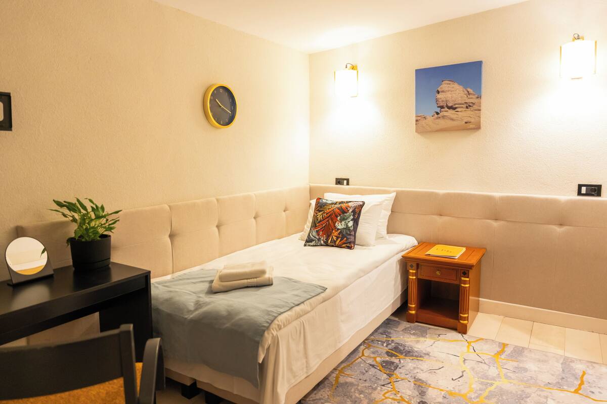 Habitación de hotel con cama, mesita de noche, lámpara, reloj y un cuadro en la pared.