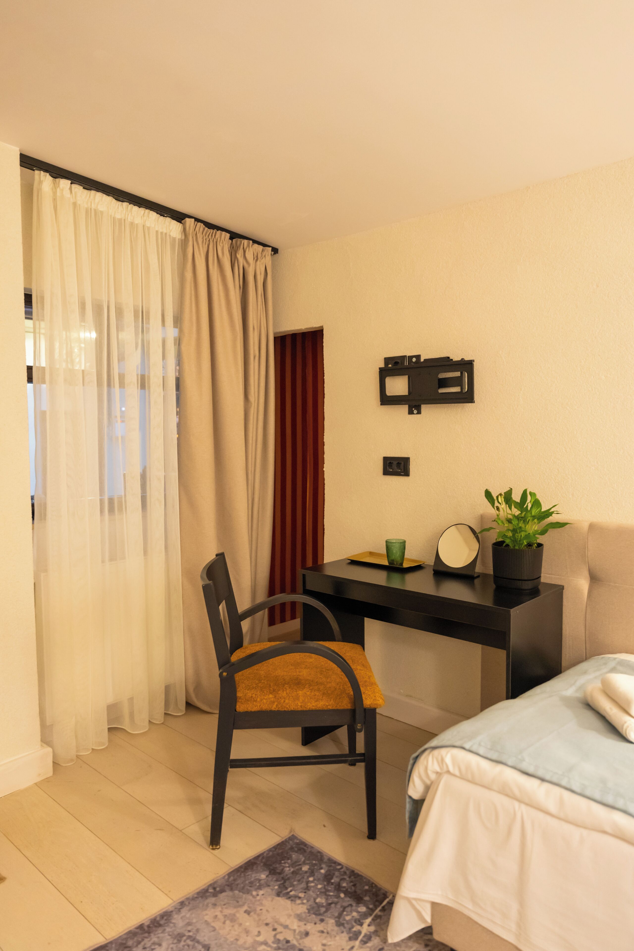 Une pièce comprenant un lit, un bureau, une chaise, une plante en pot, un miroir et un rideau.