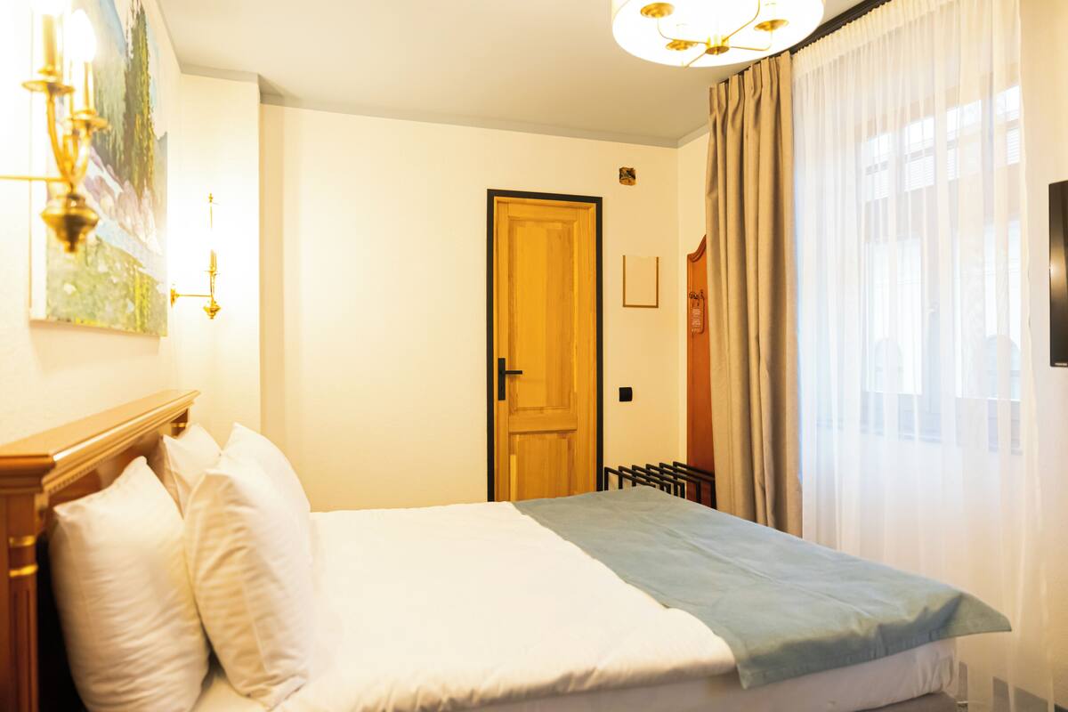 Una habitación de hotel con una cama, cabecera de madera, un cuadro en la pared, una puerta y una ventana con cortinas.