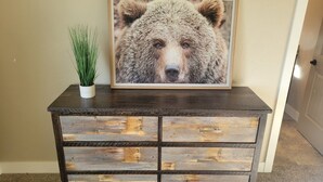 Een houten dressoir met laden tegen een muur, daarboven een lijst met een berenafbeelding, en een kleine potplant erop.