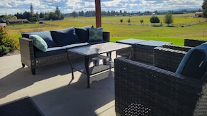 Een terras buiten met wicker meubels, een glazen tafel en uitzicht op een groen veld en verre gebouwen.