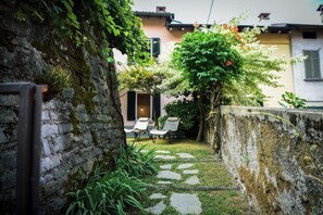 A stone pathway leading to a seating area with two chairs and a table, surrounded by greenery and a moss-covered wall. - Loppia Secret Garden by Wonderful Italy (Bellagio)
