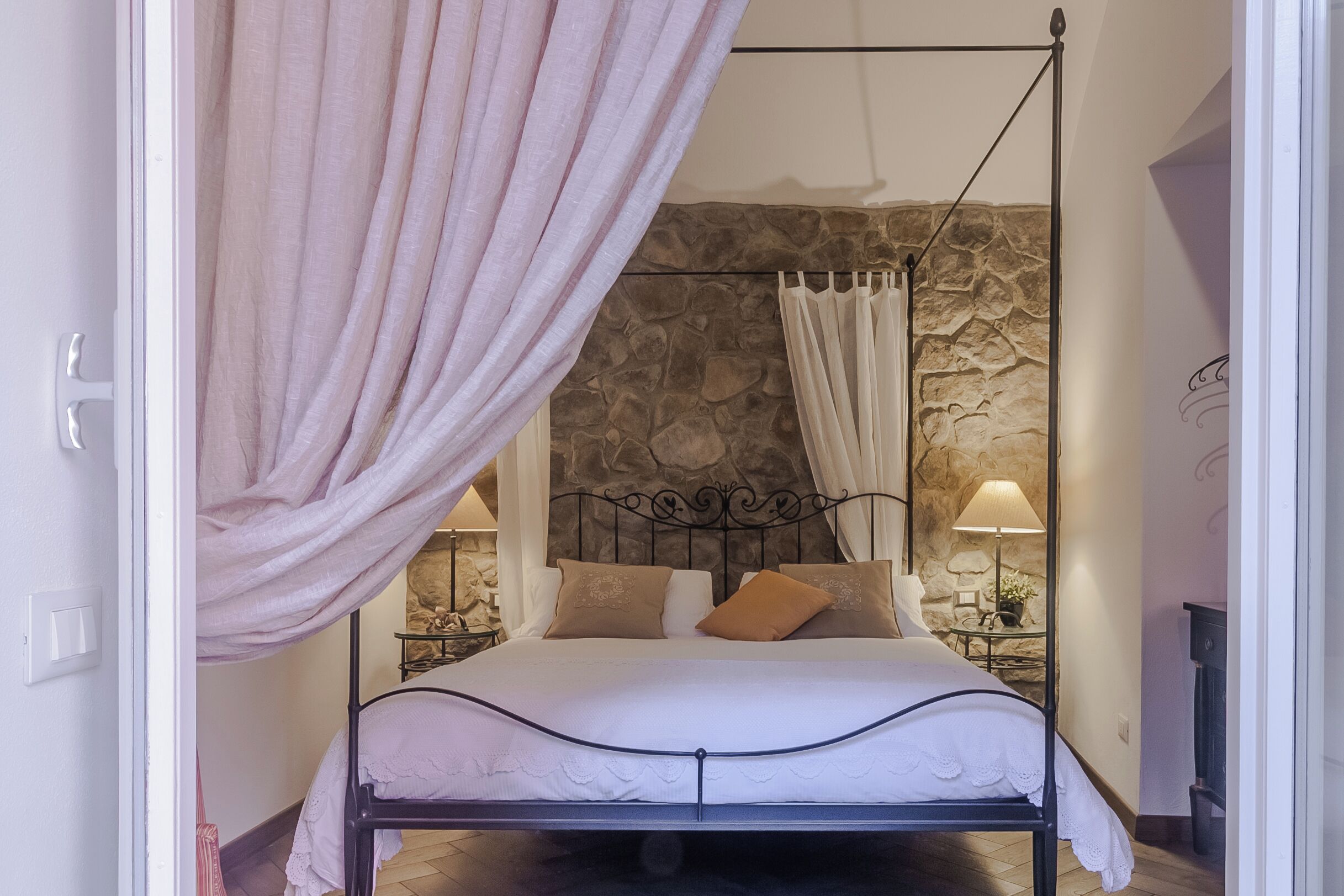 A bed with a canopy, a stone wall, and a nightstand with a lamp.