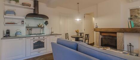 A modern kitchen with white cabinets, a built-in oven, and a gas stove. There is a blue sofa in the living area and a fireplace with a stone mantle.
