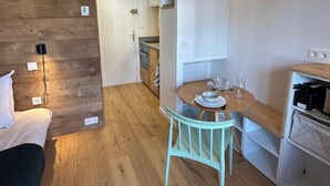 A compact room with a bed, a dining table with chairs, a kitchenette, and a wooden accent wall.