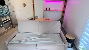 A compact living space with a sofa, a small table, a microwave, a refrigerator, and a window with blinds.