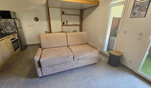 En kompakt opholdsstue med sofa, tekøkken og skydedør til terrassen.