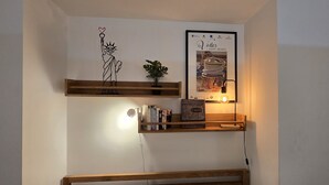 A wall with two wooden shelves, one holding books and a small plant, and the other displaying a framed poster.