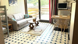 A living room with a sofa, coffee table, and a TV. There is a window with curtains, a radiator, and a view of a forest. - Mountain Apartment With Terrace (Saint-Chaffrey)