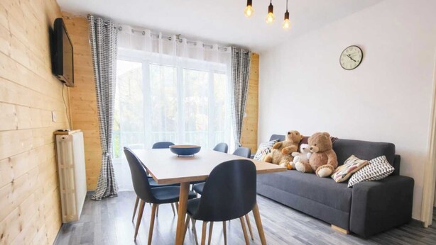 A cozy room with a dining table, chairs, a sofa with teddy bears, a clock, and a window with curtains.