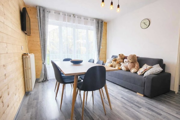 A cozy room with a dining table, chairs, a sofa with teddy bears, a clock, and a window with curtains.