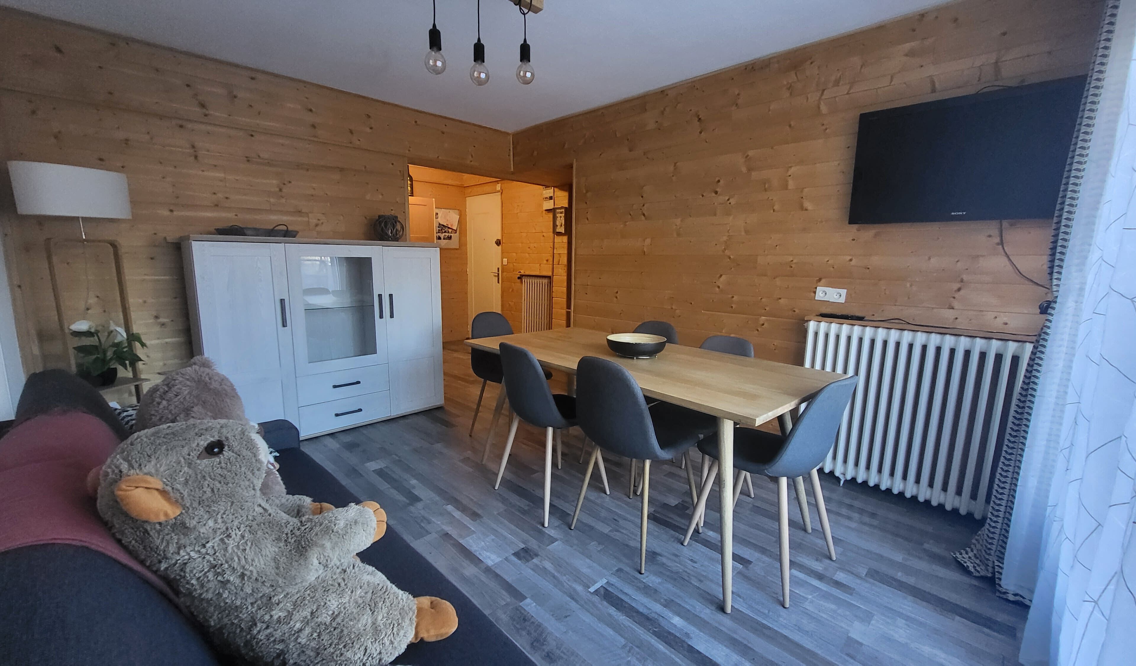 Een knus vertrek met houten inrichting, een eettafel met stoelen, een bank met pluchen speelgoed en een aan de muur bevestigde televisie.
