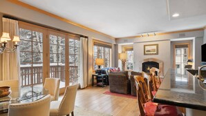 A spacious living area with a fireplace, a dining table with chairs, and large windows offering a view of the outdoors.