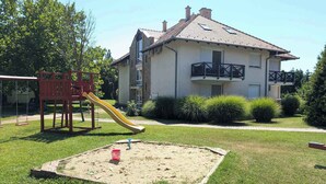 Un parque infantil con tobogán y columpios, un arenero y un edificio de dos plantas con balcones.