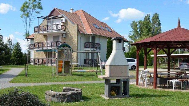 Un edificio con techo de tejas rojas, una zona de barbacoa con chimenea y un porche con mesas y sillas.