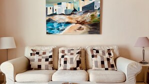 A living room with a beige sofa, two patterned throw pillows, a white coffee table with a decorative plate, and a painting of a coastal scene above the sofa.
