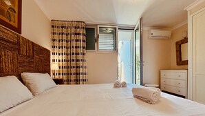 A bedroom with a large bed, a nightstand with a lamp, a dresser, and a window with blinds.