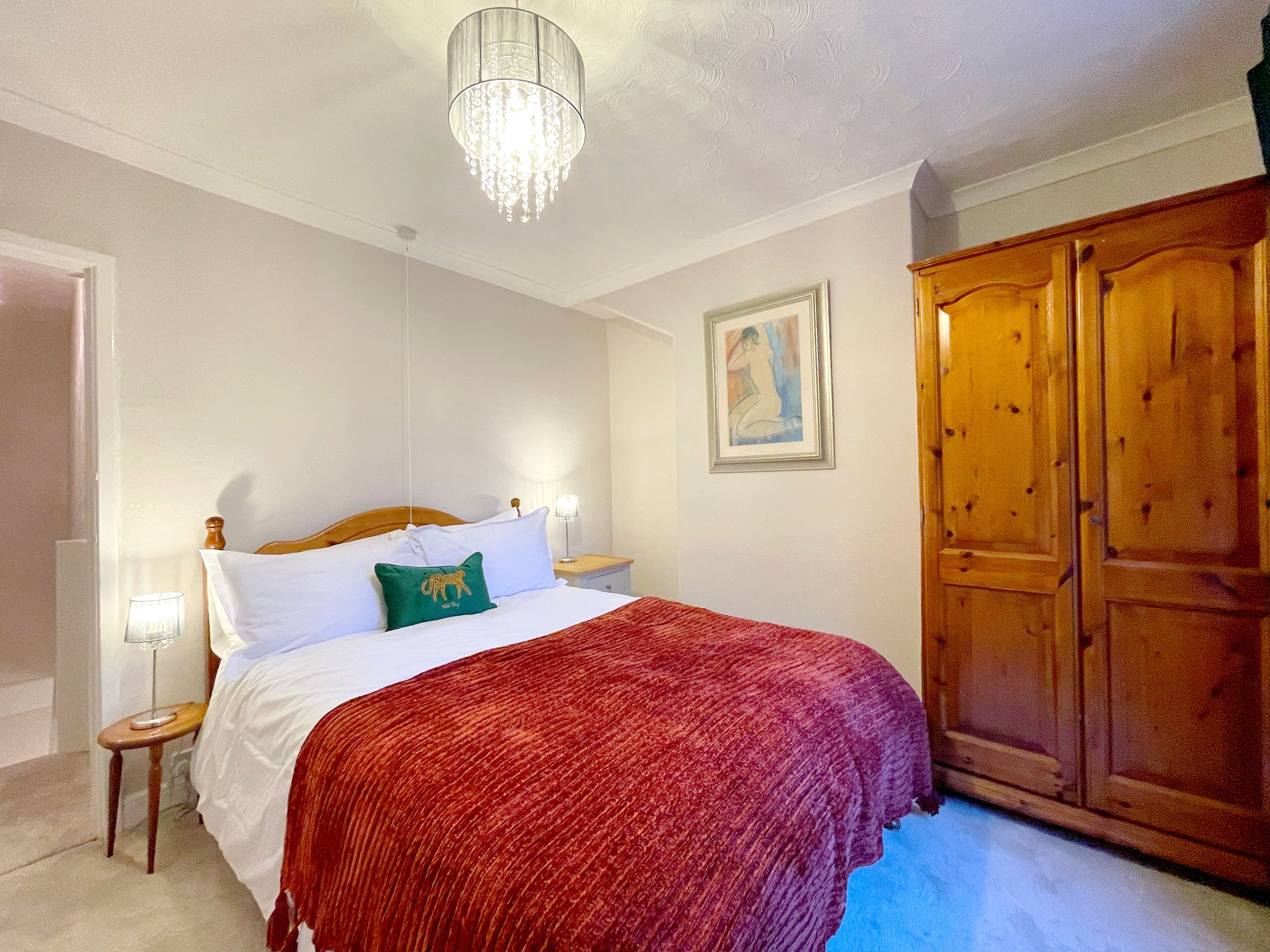 Een slaapkamer met een houten garderobe, een bed met witte lakens en een rode deken, een kroonluchter en een lijstwerk met kunst aan de muur.