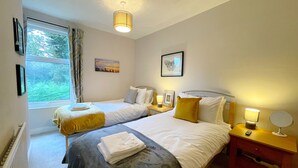 A hotel room with two beds, a window with curtains, a bedside table with a lamp, and a wall-mounted picture. - Stylish cottage in vibrant riverside village (North Ferriby)