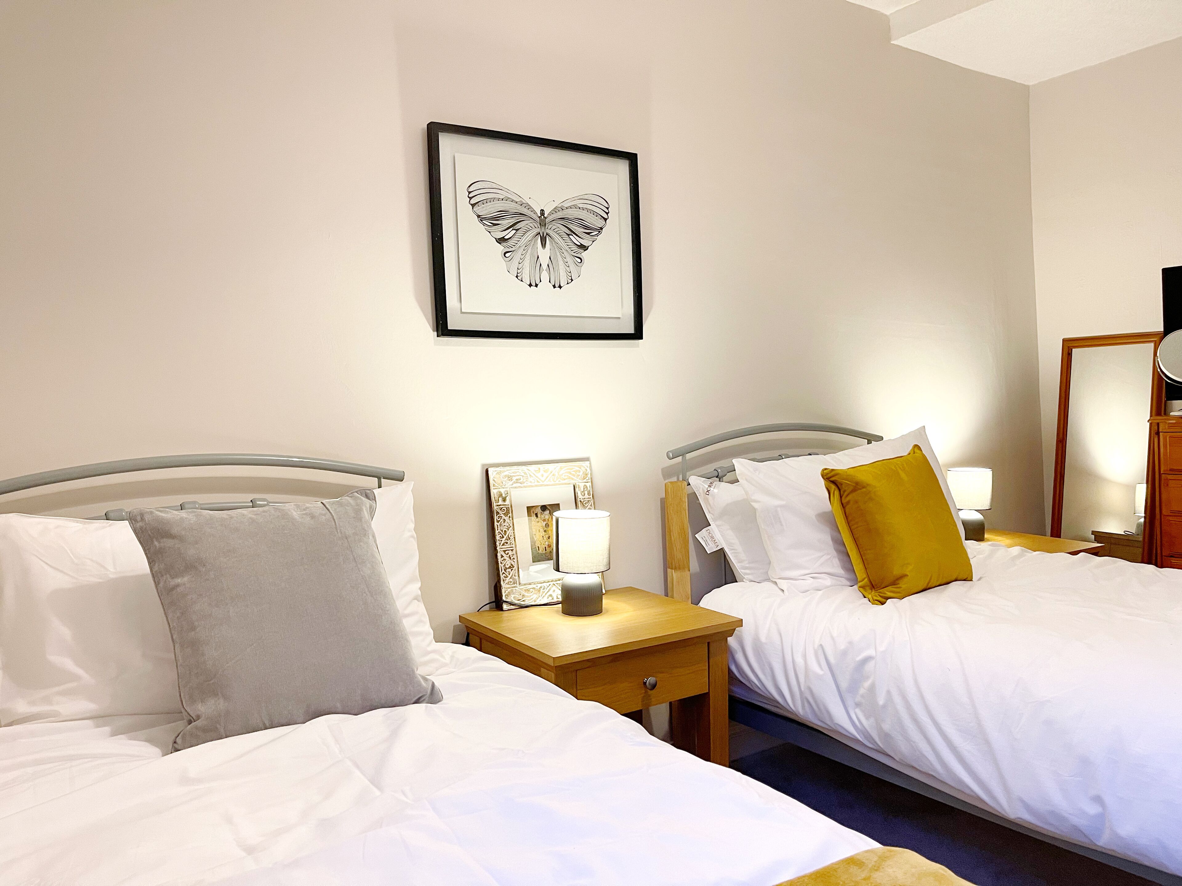 A hotel room with two beds, each with white bedding and pillows, a bedside table with a lamp, and a framed butterfly artwork on the wall.