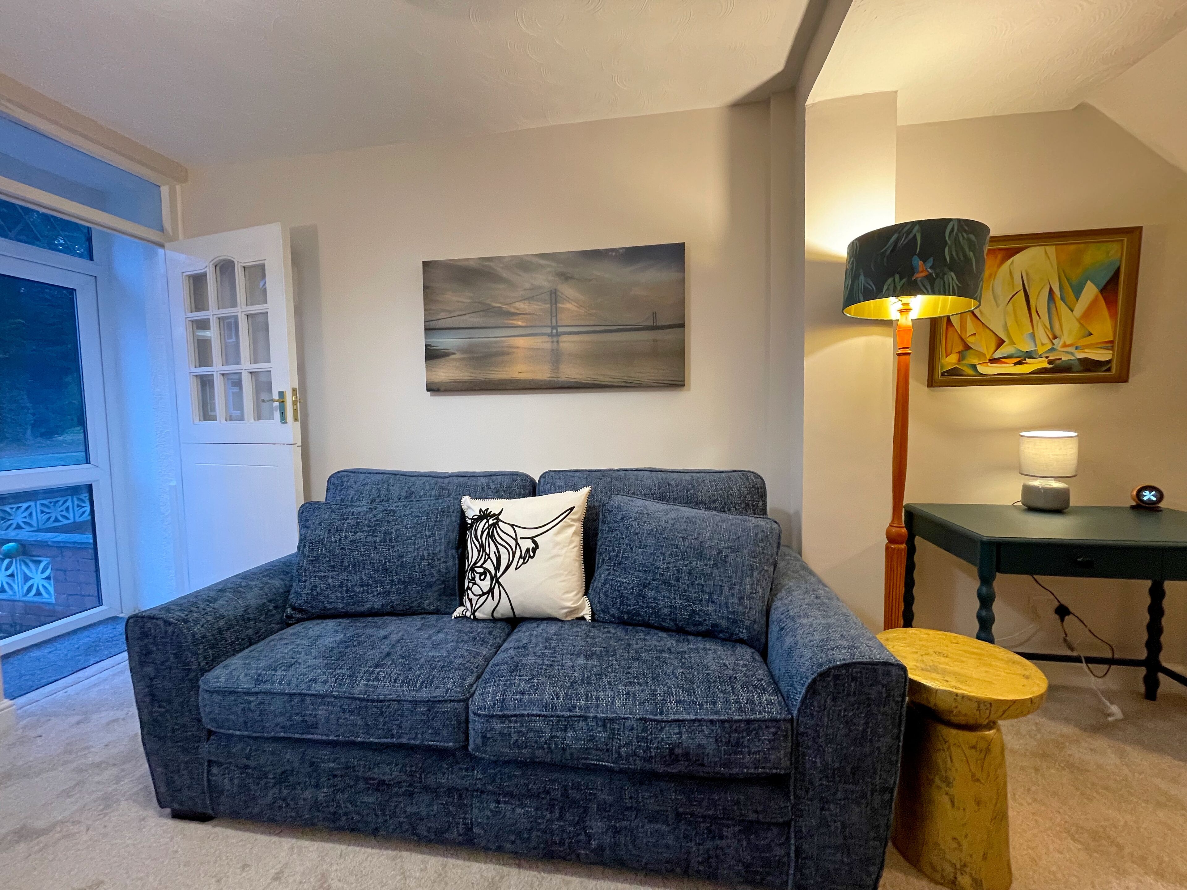 Una sala de estar con un sofá azul, un cuadro, una lámpara y una mesa.