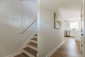 Un escalier avec des marches en bois et des murs blancs, menant à un espace cuisine avec des armoires blanches et une fenêtre.