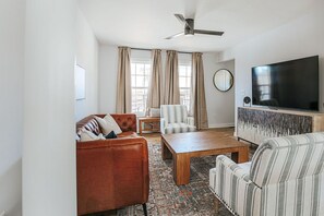 Een woonkamer met een bruine leren bank, een gestreept fauteuil, een houten salontafel, een televisie op een witte kast en een plafondventilator.