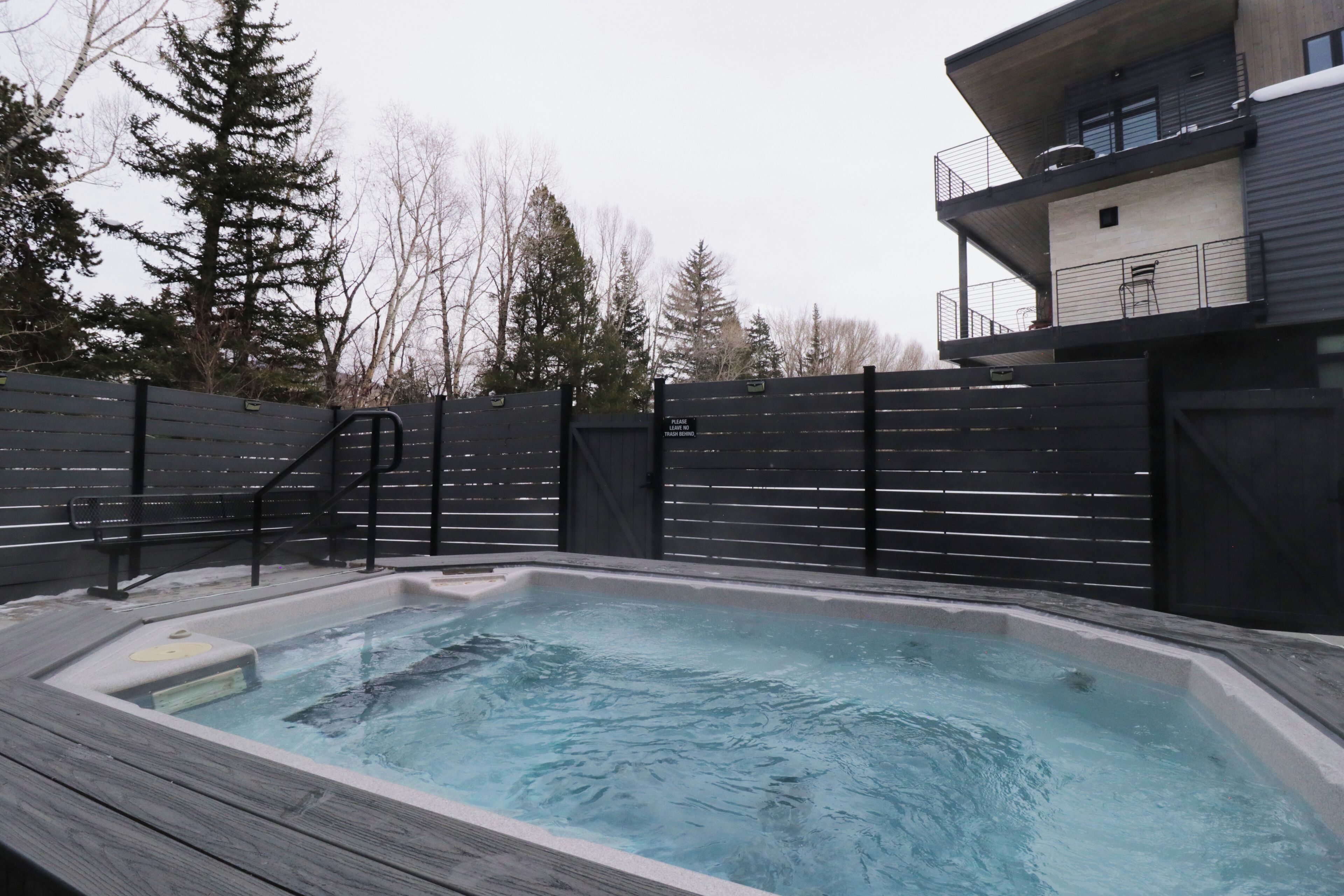 Ein Whirlpool mit einer Holzterrasse, umgeben von einem schwarzen Zaun, und ein Gebäude mit Balkonen im Hintergrund.
