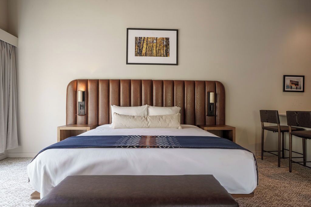 A hotel room with a bed, a bedside table, a chair, a bench, and a framed picture on the wall.