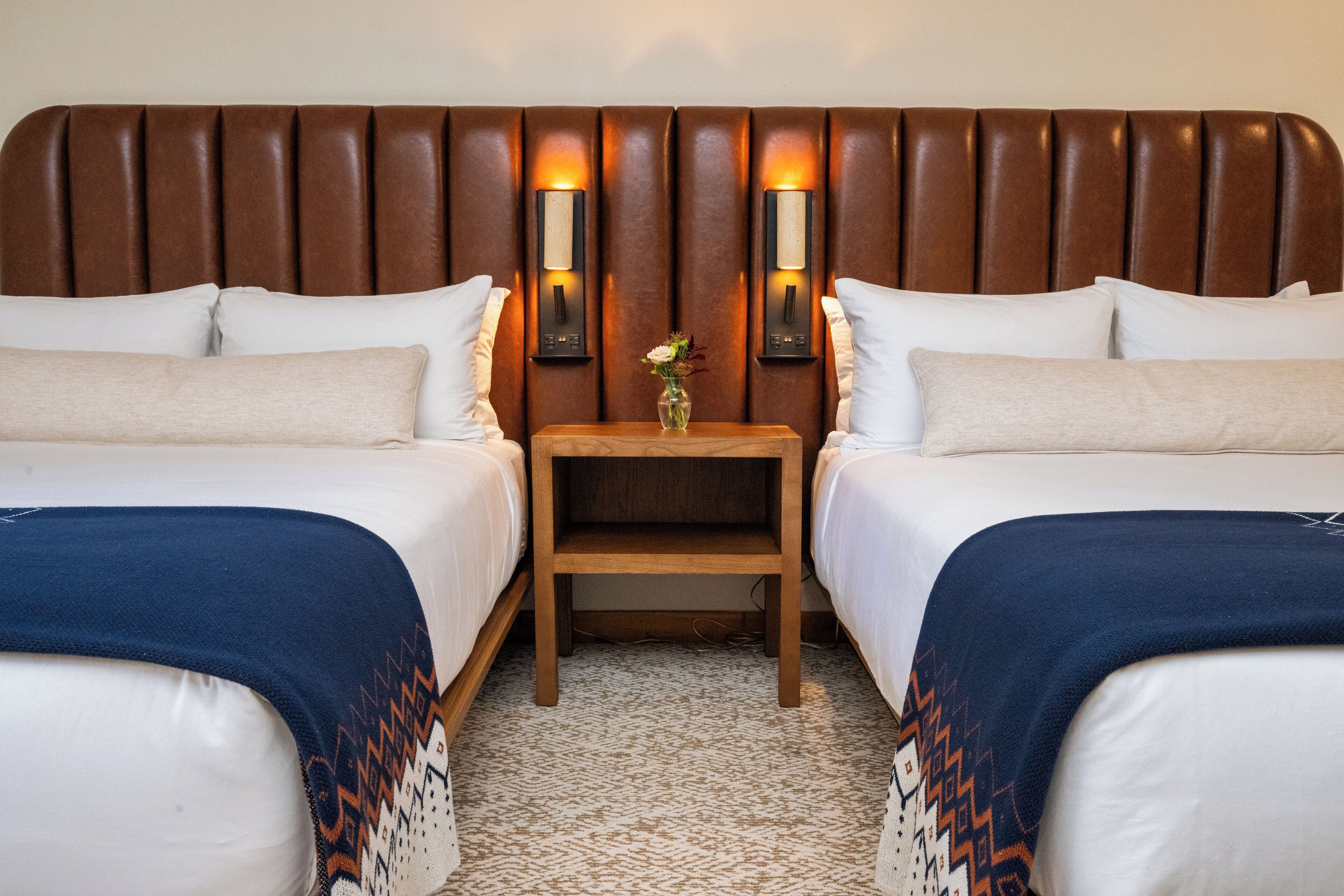 A hotel room with two beds, a wooden nightstand with a vase, and wall-mounted lamps.