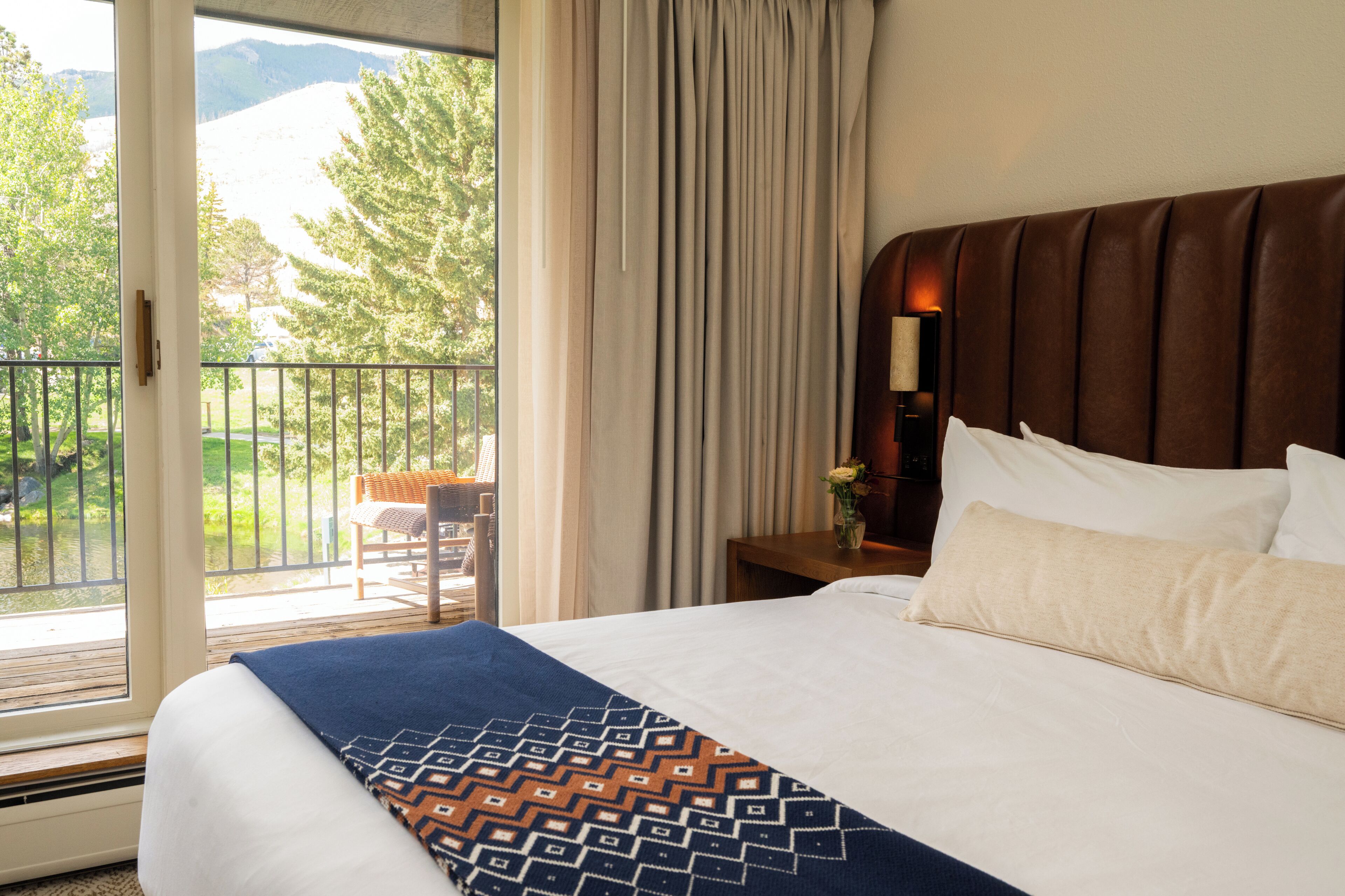 A hotel room with a large bed, a wooden headboard, a nightstand with a lamp, a window with curtains, and a balcony with a chair.