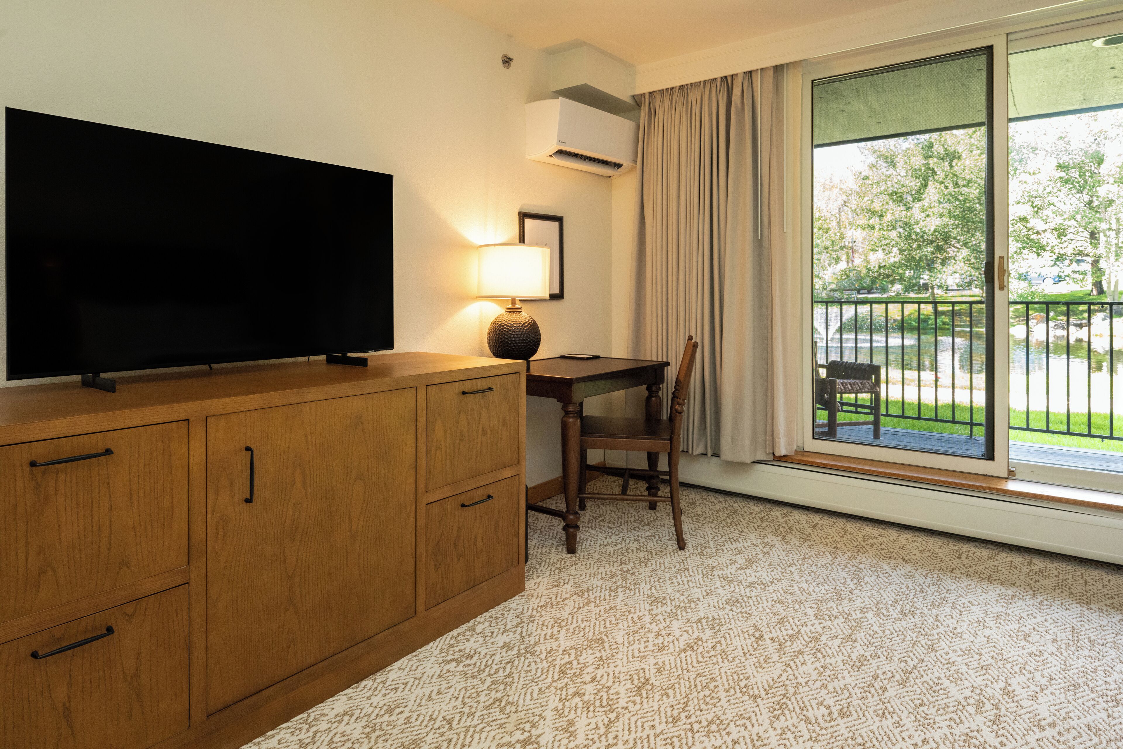 A hotel room with a wooden cabinet, a flat-screen TV, a table with a chair, a lamp, and a large window with a view of a garden and a body of water.