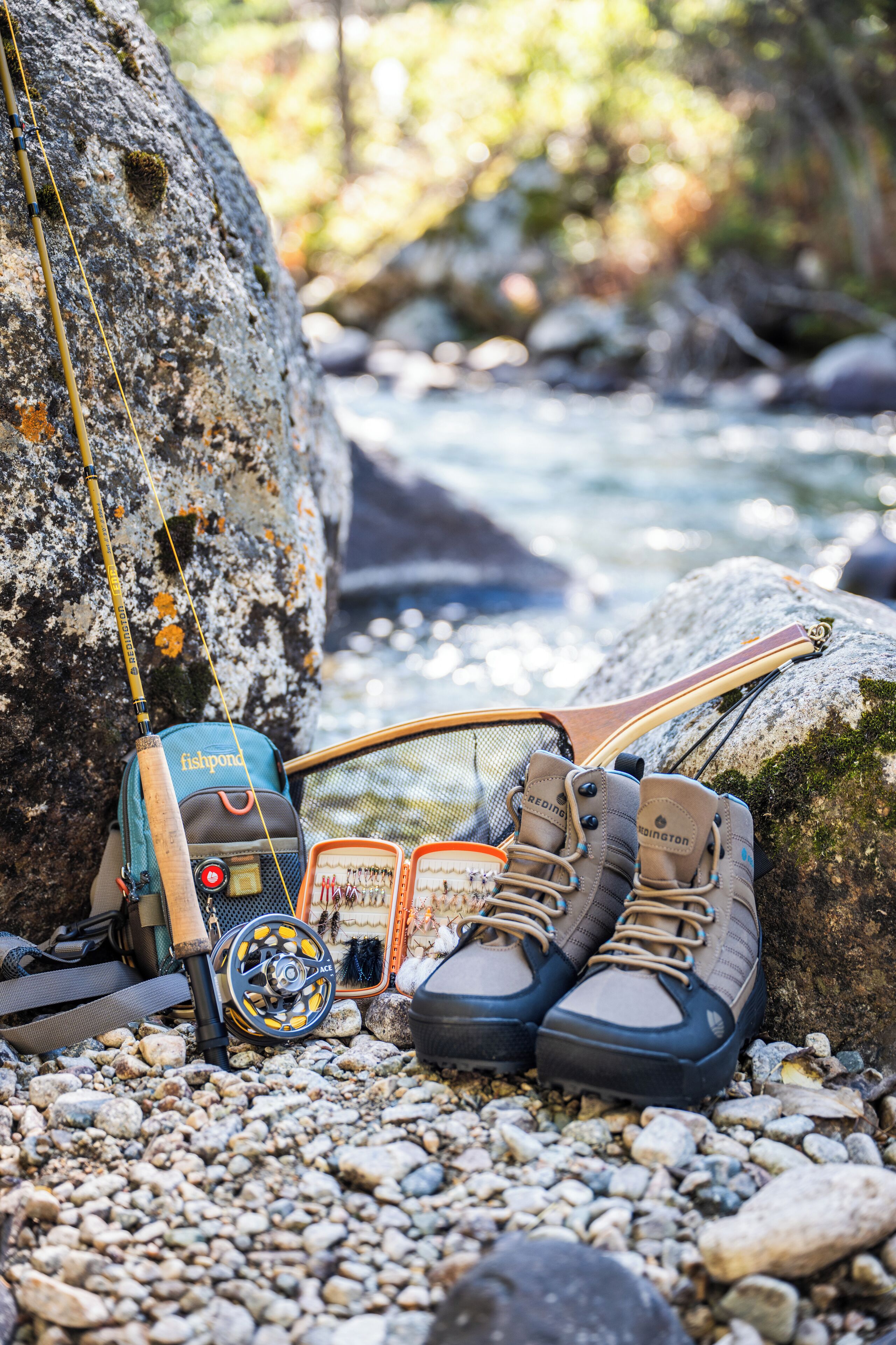 A fishing rod, a fishpond, and a pair of hiking boots on a rocky riverbank.