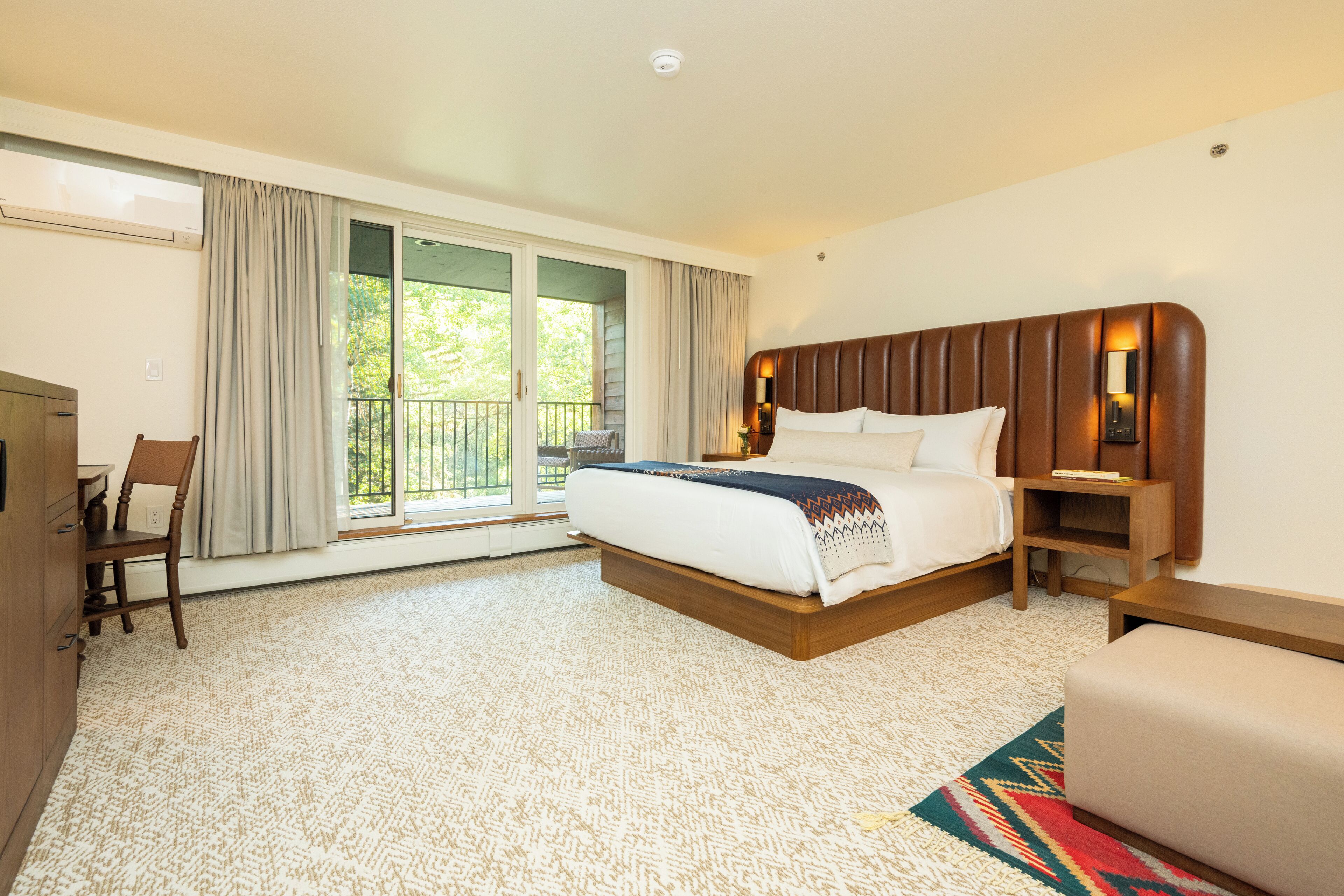 A hotel room with a large bed, a desk, and a chair. There is a balcony with curtains and a view of greenery.