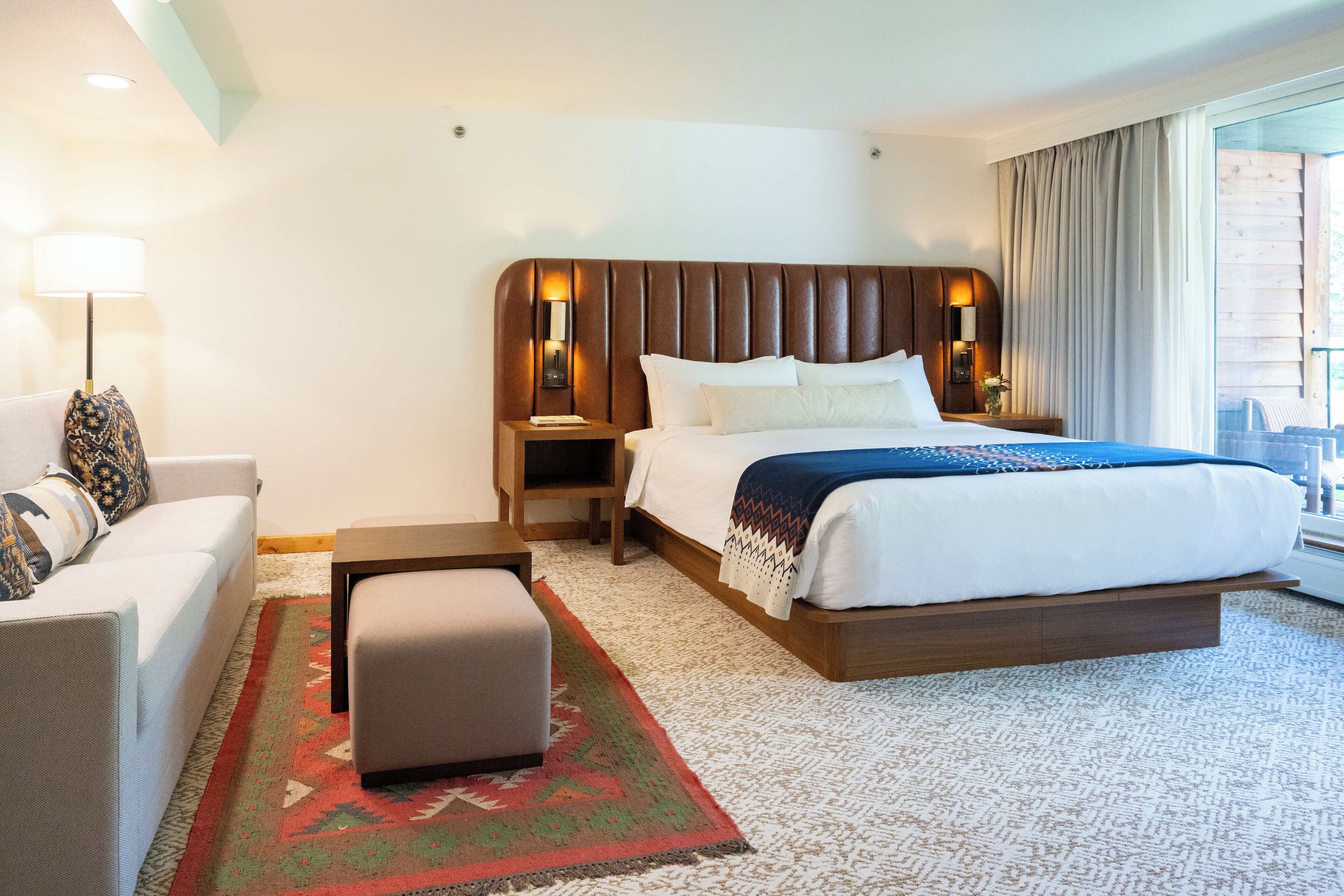 A hotel room with a large bed, a nightstand, a lamp, a sofa, and a view of the city.