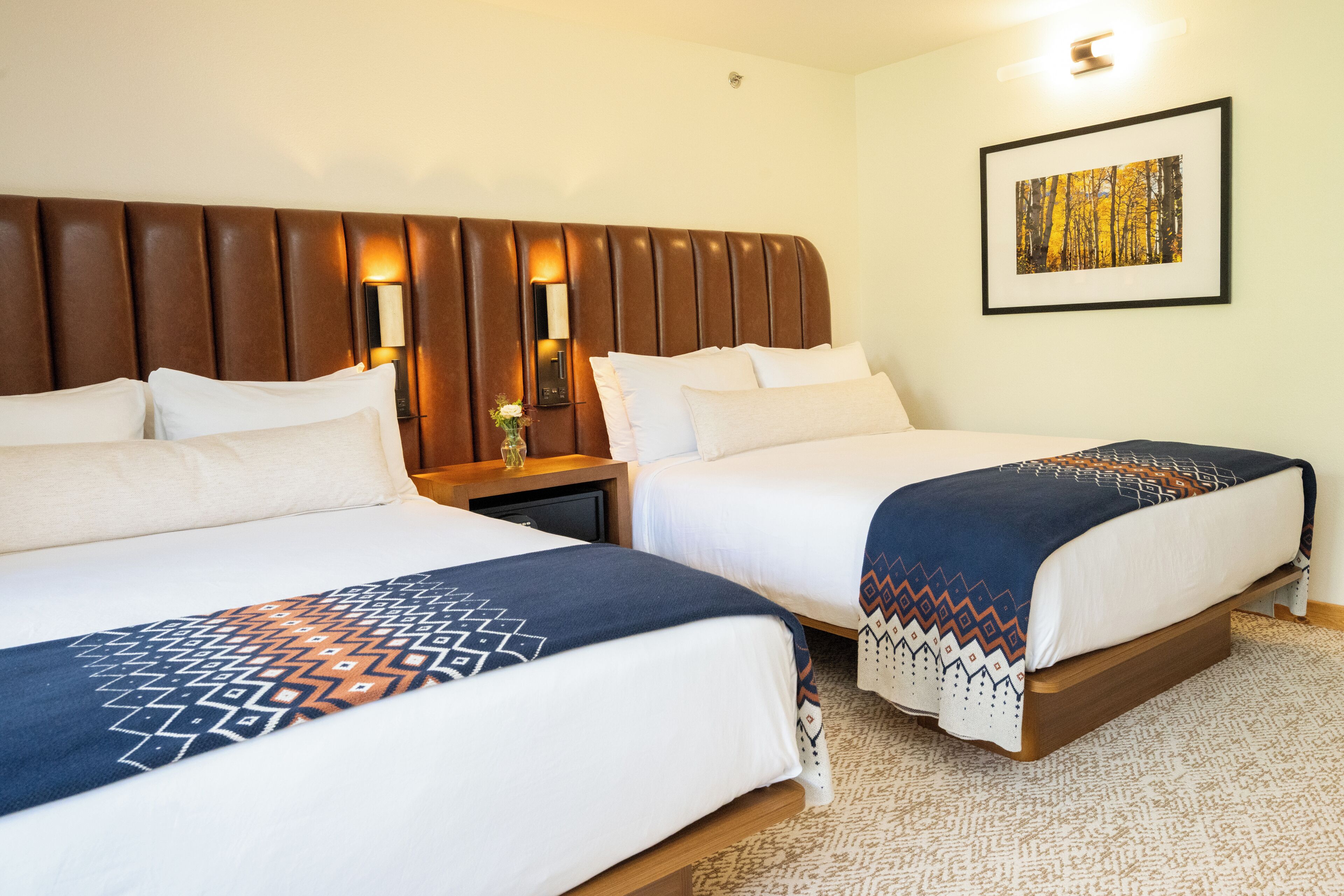 A hotel room with two beds, a wooden headboard, a nightstand with a lamp, and a framed picture on the wall.