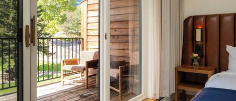 A hotel room with a balcony, wooden flooring, a bed, a nightstand, and a chair.